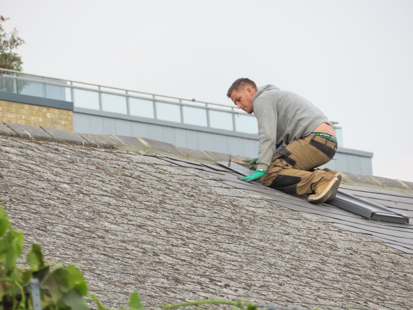 A man in a gray hooded sweatshirt kneeling on a sloped roof, hammering a course of dark shingles into place under an overcast sky