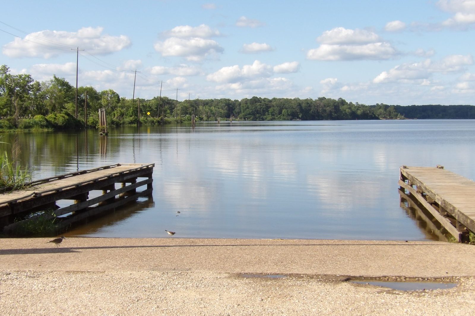 An empty concrete boat ramp running into a flat calm lake between two low wooden courtesy docks, with a wooded far shore under a partly cloudy sky