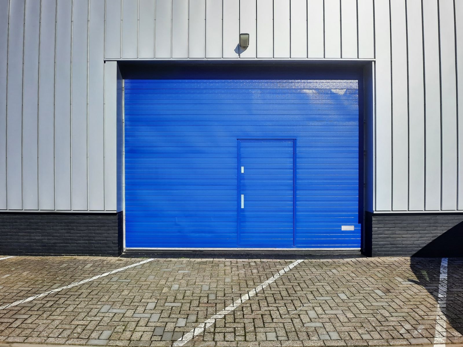 A bright blue roll-up loading door set into the metal-clad wall of a commercial building, with paved parking in front