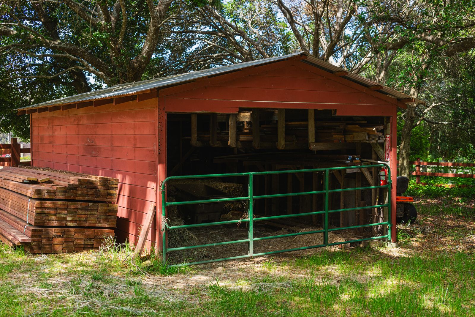 A red outbuilding with an open bay, stacked lumber, and a metal gate on a rural property