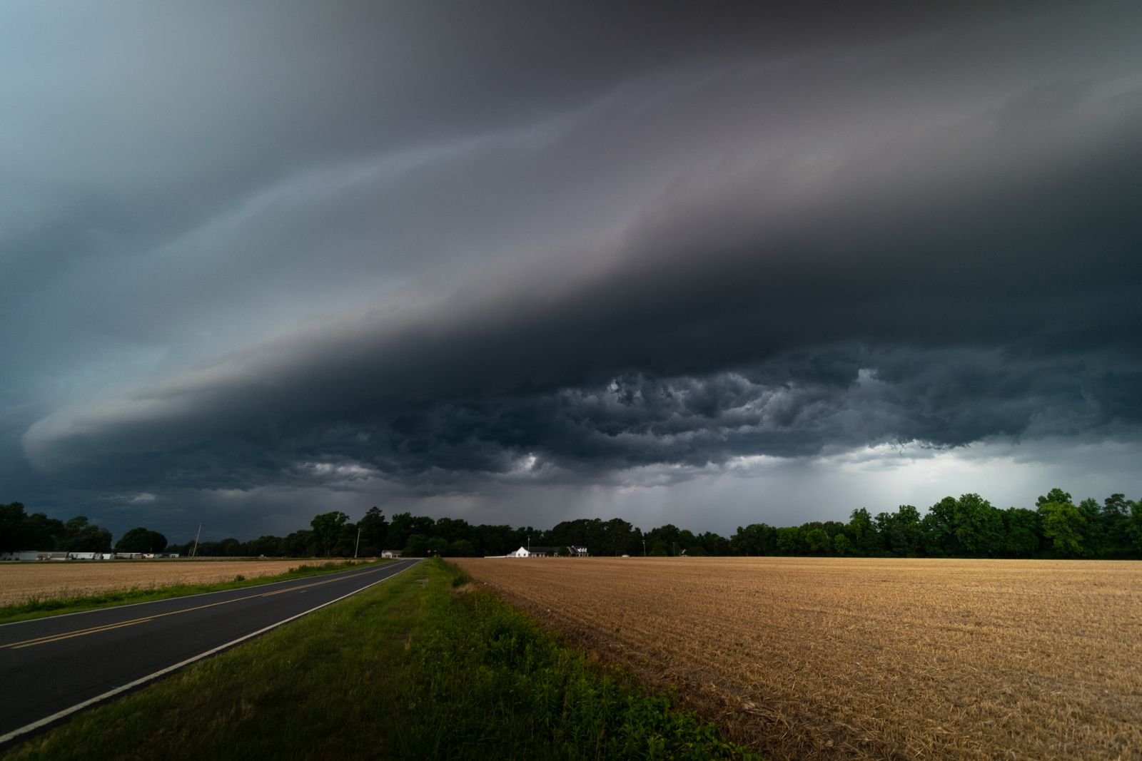 A dark shelf cloud rolling over a two-lane country road and a harvested field, with a tree line and a white farmhouse under the storm