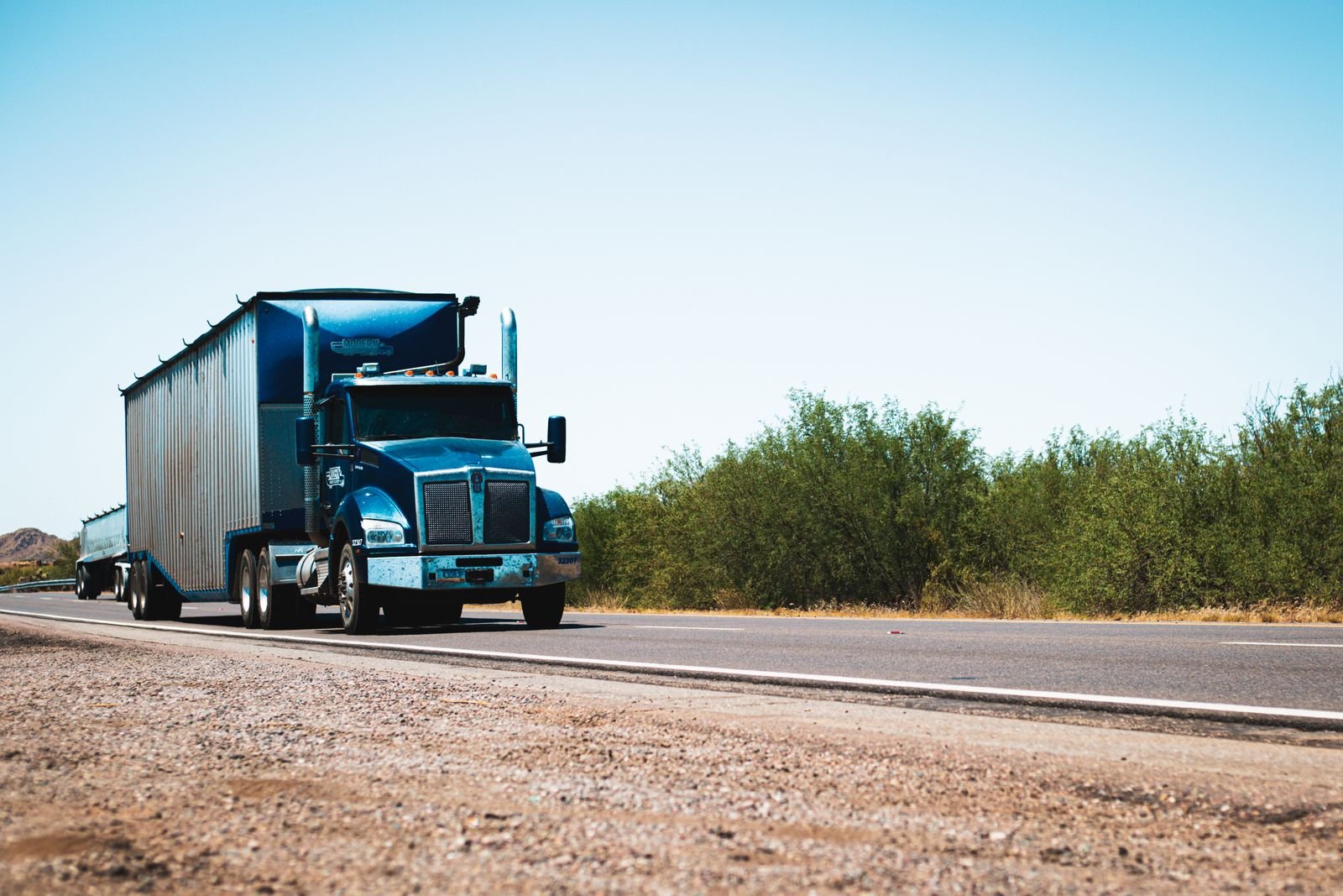 A blue semi truck pulling a dry van trailer down a rural two-lane highway lined with green brush under a clear sky