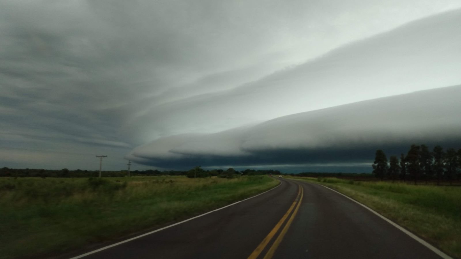 A dark shelf cloud rolling in low over an empty two-lane highway with a yellow center line, flat farmland and a tree line on either side