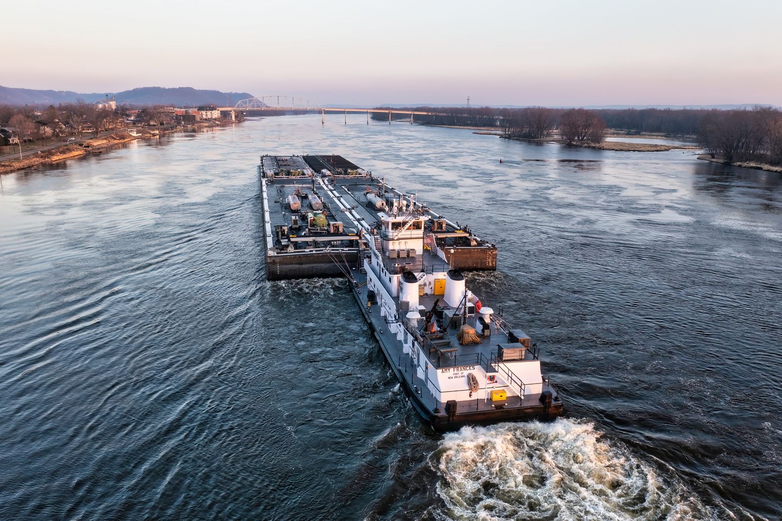 A towboat pushing a string of loaded barges down a wide river at dusk, with low wooded banks, bare winter trees and a distant bridge