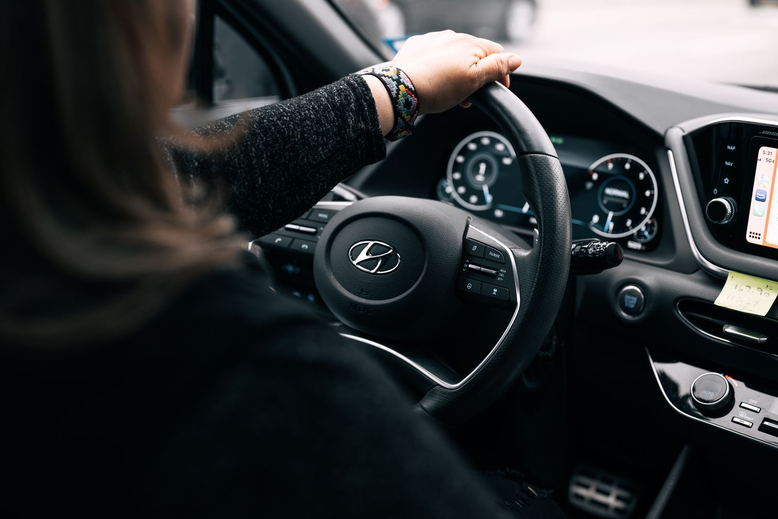 Over-the-shoulder view of a driver's hand resting on a car steering wheel, with the instrument cluster and infotainment screen lit up beyond it