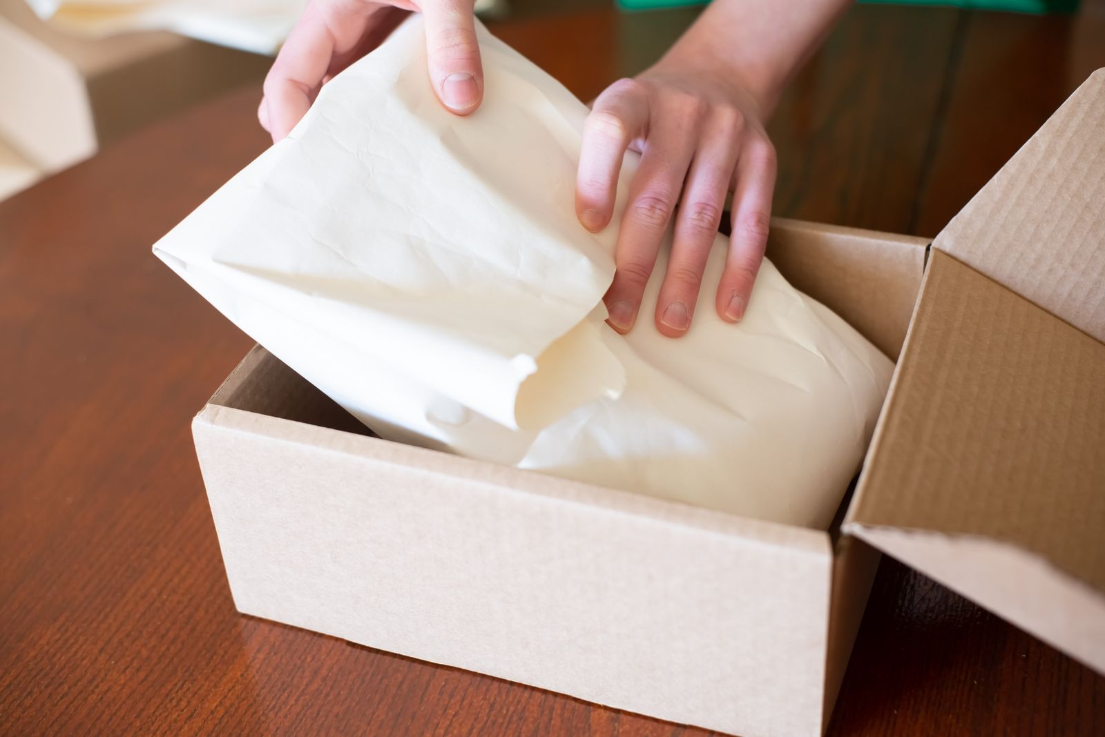 Two hands wrapping a small item in white tissue paper and settling it into an open cardboard shipping box on a dark wooden table
