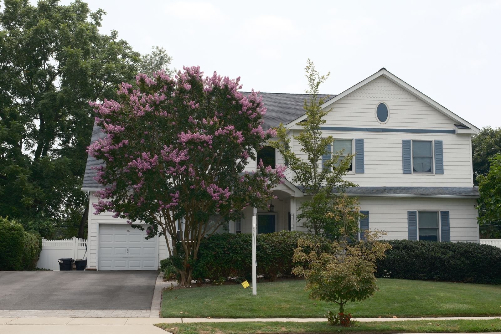 A two-story white house with black shutters and an attached garage, a pink crape myrtle blooming in the front yard beside the driveway