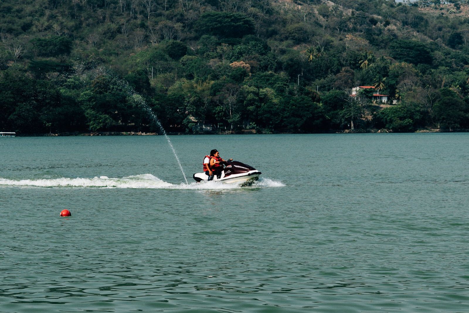 A rider in a life jacket on a personal watercraft crossing a tree-lined lake