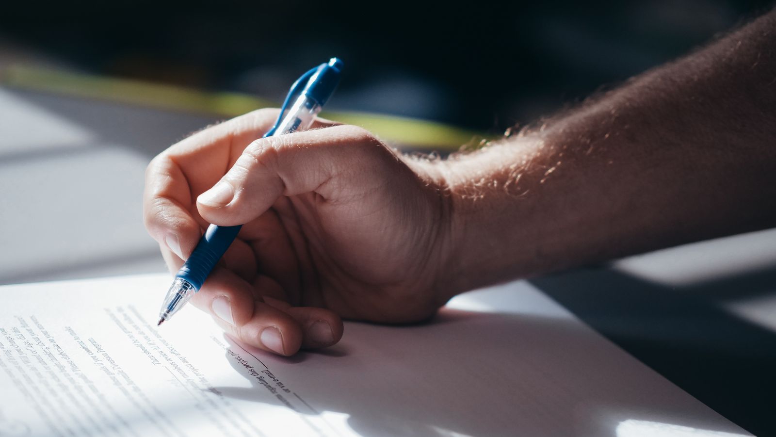 A hand holding a blue ballpoint pen, poised over a printed form on a desk in low, angled light