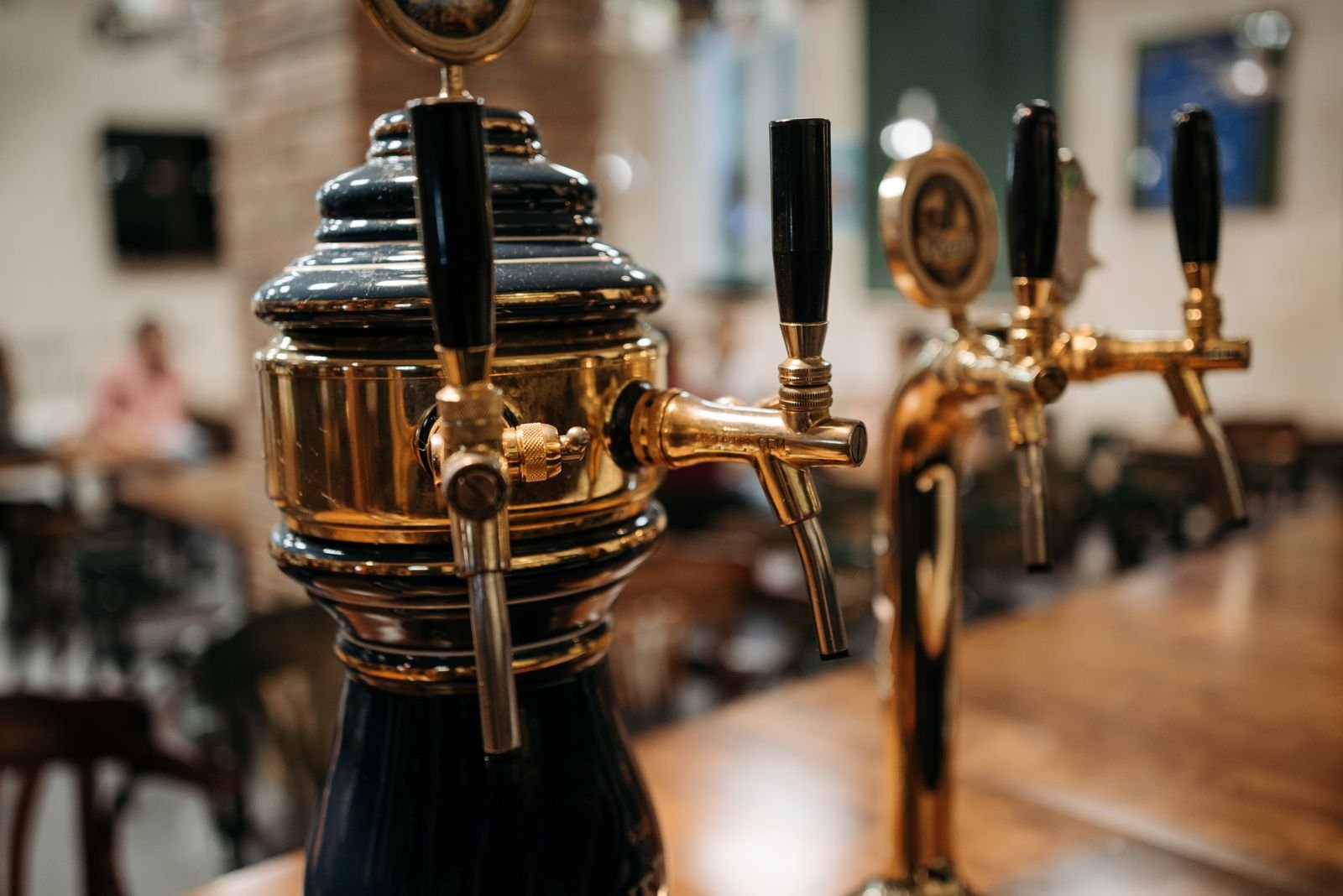 Polished brass beer taps on a wooden bar counter, with the dim interior of a bar blurred in the background