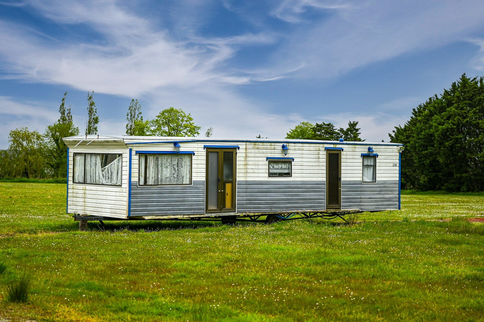 A white and blue single-wide mobile home resting on its steel chassis in a green field under a blue sky