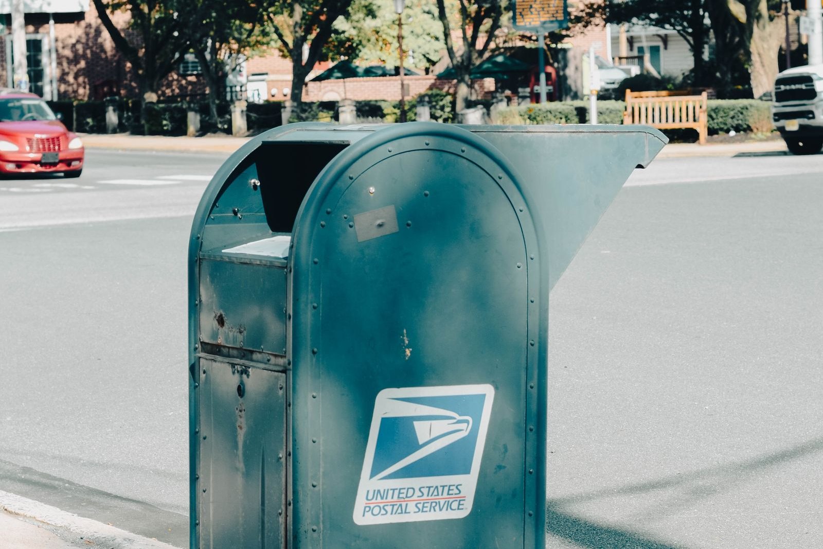 A weathered blue United States Postal Service collection box standing on a curb along a tree-lined street