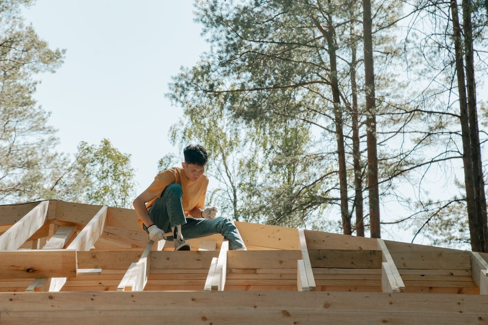 A worker in a yellow shirt kneeling on the new wood roof framing of a house under construction, with pine trees behind him
