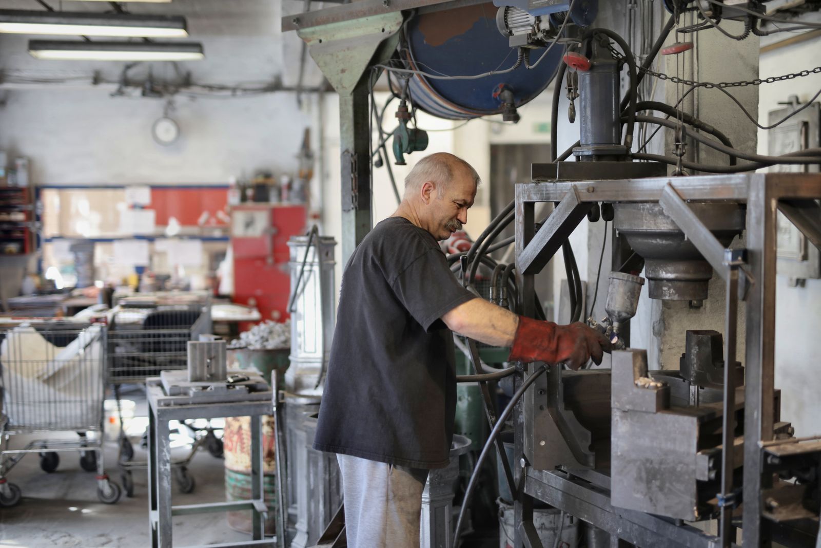 A gray-haired man in a dark T-shirt and a single red work glove operating a press in a small, cluttered metal shop