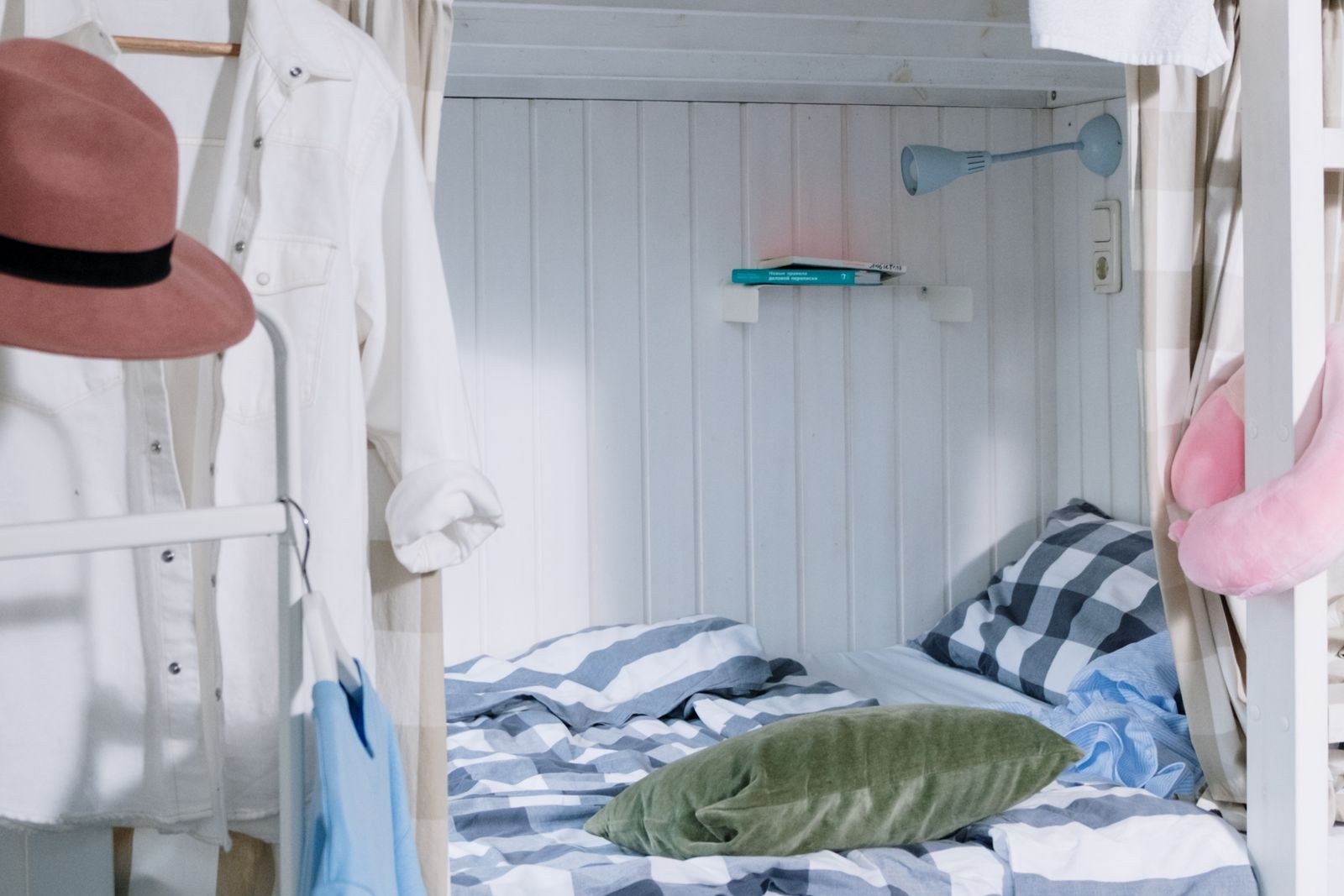 The lower bunk of a bed in a white-paneled shared room, with checked bedding, a white shirt and a pink hat hanging beside it, and two books on a small wall shelf