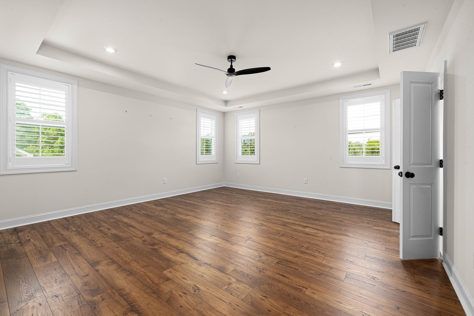 An empty room in a house with bare hardwood floors, white walls, a ceiling fan, and daylight coming through four windows