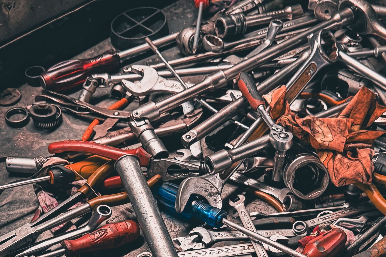 A cluttered metal toolbox tray piled with wrenches, sockets, ratchets, pliers and screwdrivers beside a work glove
