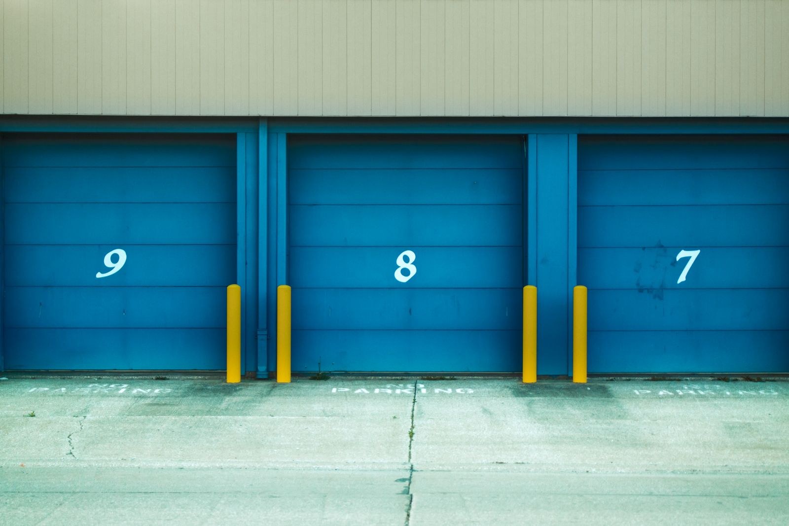 Three blue roll-up storage unit doors numbered 9, 8 and 7, with yellow bollards along a concrete drive